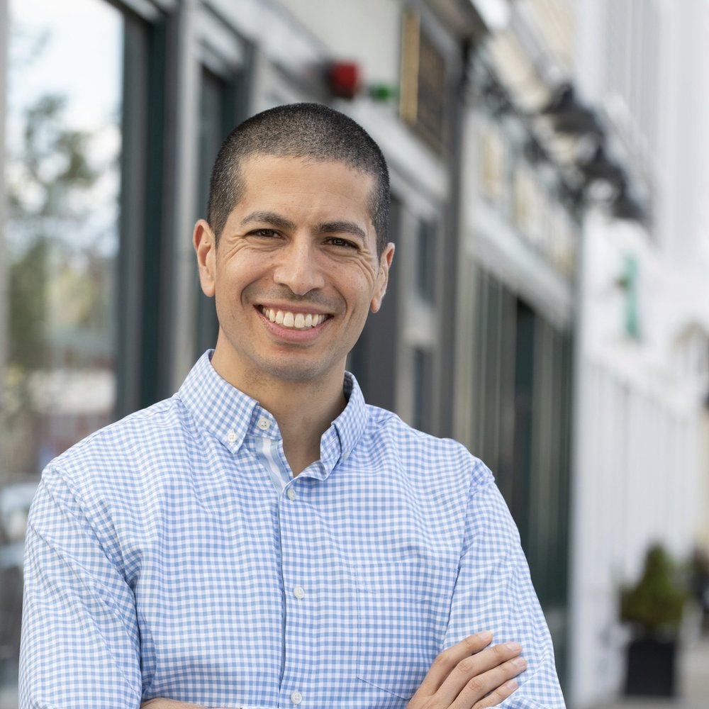 Dan Koh, the Democratic nominee, in a checked shirt with arms crossed on a storefront sidewalk
